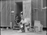 3 July 1942. Evacuees family of Japanese ancestry relax in front of their barrack room at the end of day. The father is a worker on the farm project at this War Relocation Authority center. Note the chair which was made of scrap lumber, and the wooden shoes known as Getas made by evacuees. National Archives, 537989.